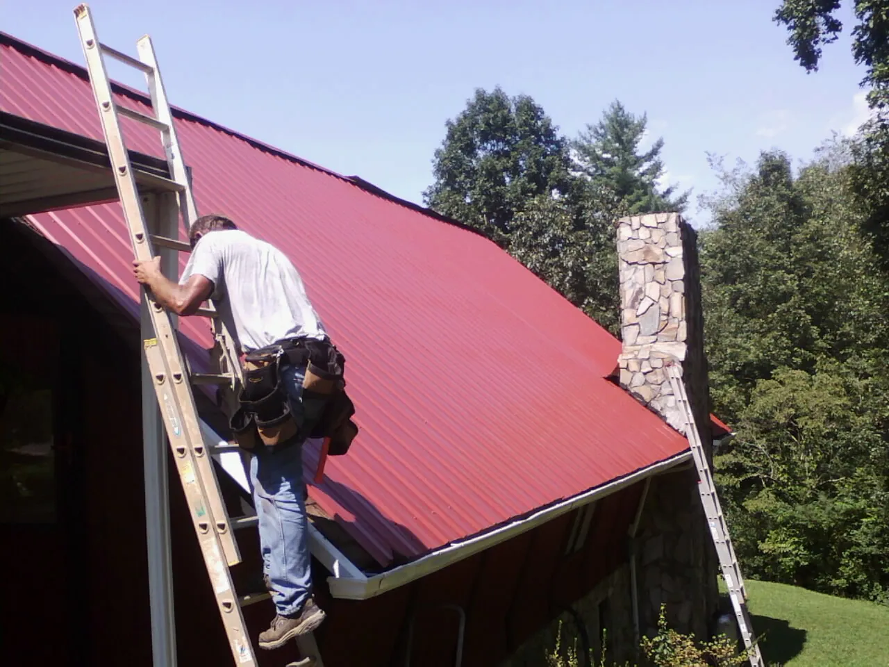 Roofer on a ladder beside a red metal roof and stone chimney — Soffit & Fascia Repair by Roofing Brothers in Ronks